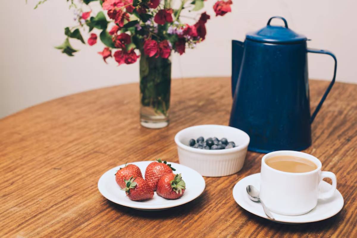 A rustic wooden table set with fresh strawberries, blueberries, a cup of coffee, and a blue teapot, alongside a colorful flower vase.