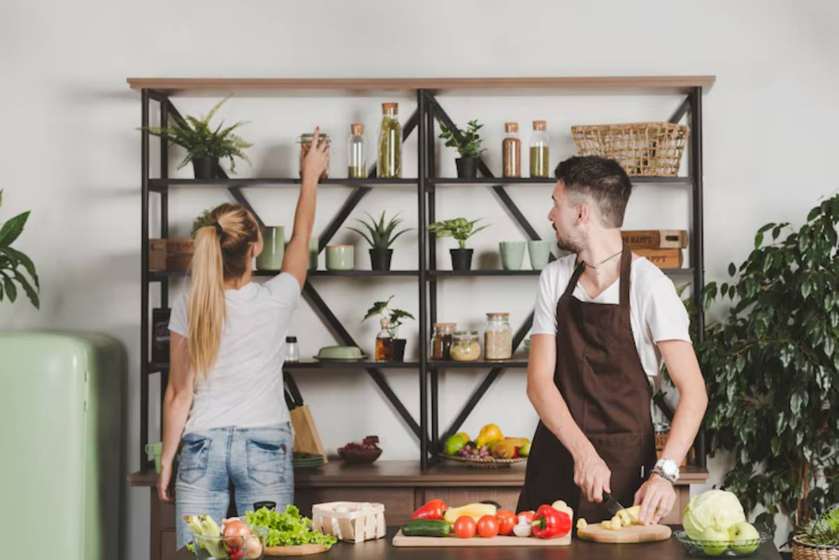 A modern kitchen scene with a man chopping vegetables on a wooden table while a woman reaches for items on a shelf. Green plants are present.