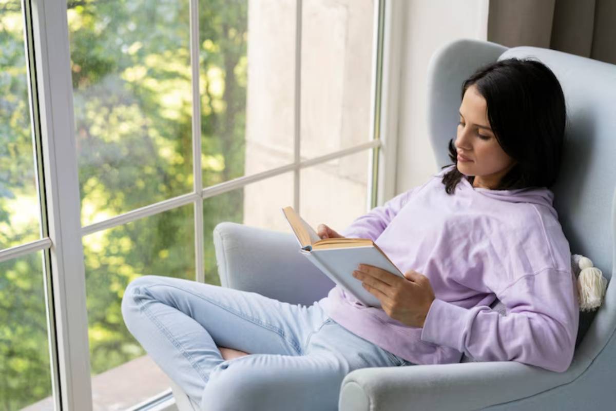 A person relaxes in a cozy chair by a large window, reading a book with feet propped up, surrounded by greenery.