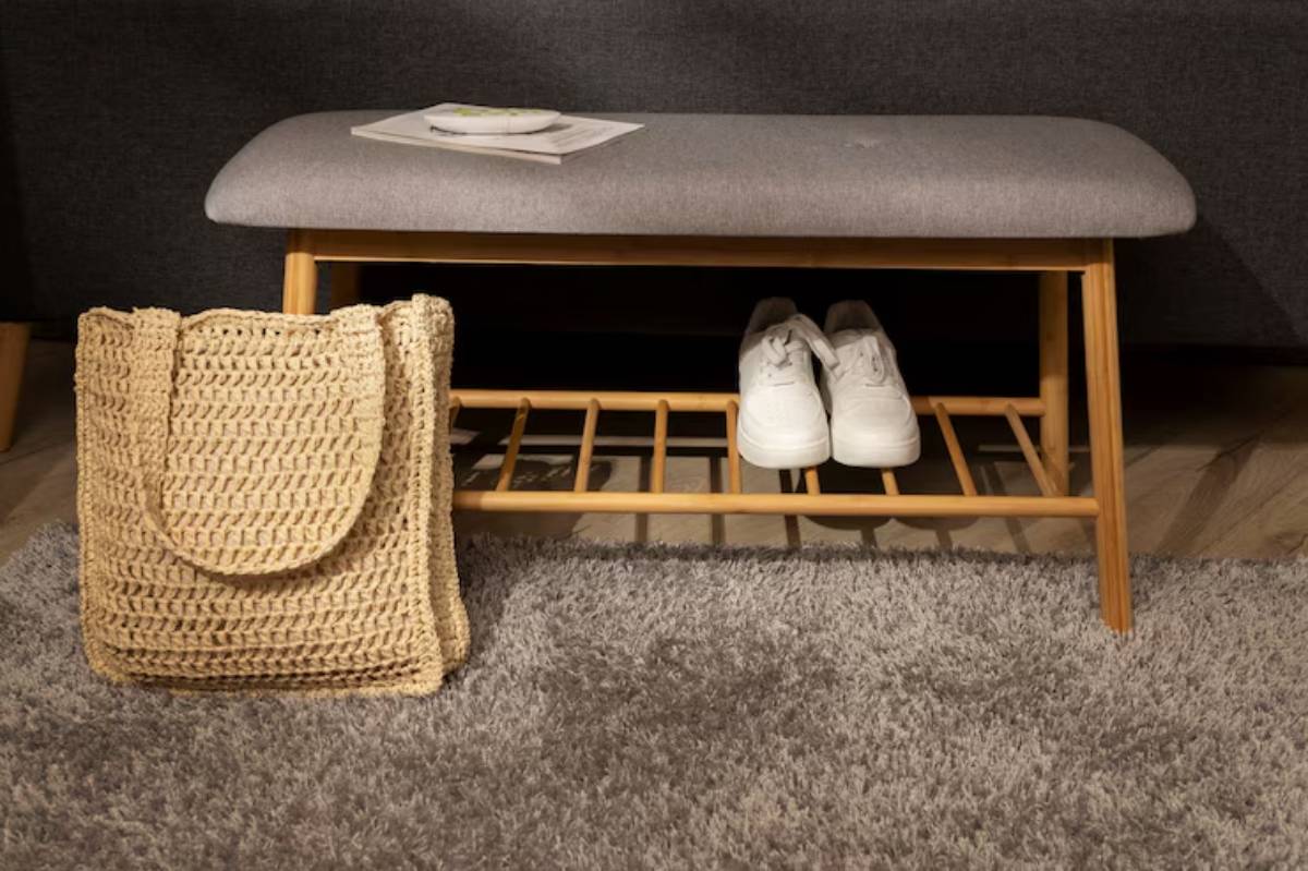 A wooden bench with a gray cushion, a woven tote bag, and white sneakers stored underneath, placed on a soft rug.