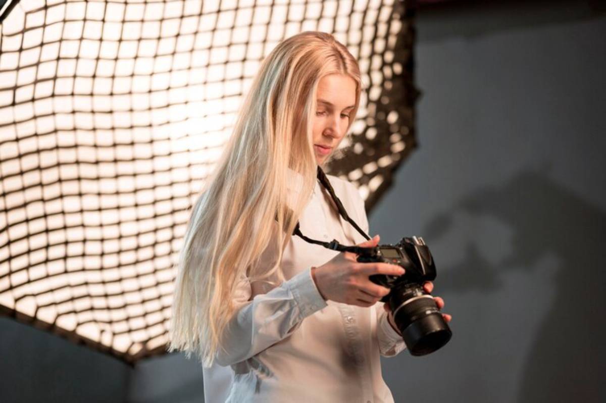 A person in a white shirt holds a camera, standing in front of a large softbox, with soft lighting creating a studio ambiance.
