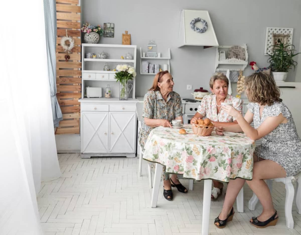 Three women enjoy tea and pastries at a floral table in a cozy, bright room with light wood and soft decor.