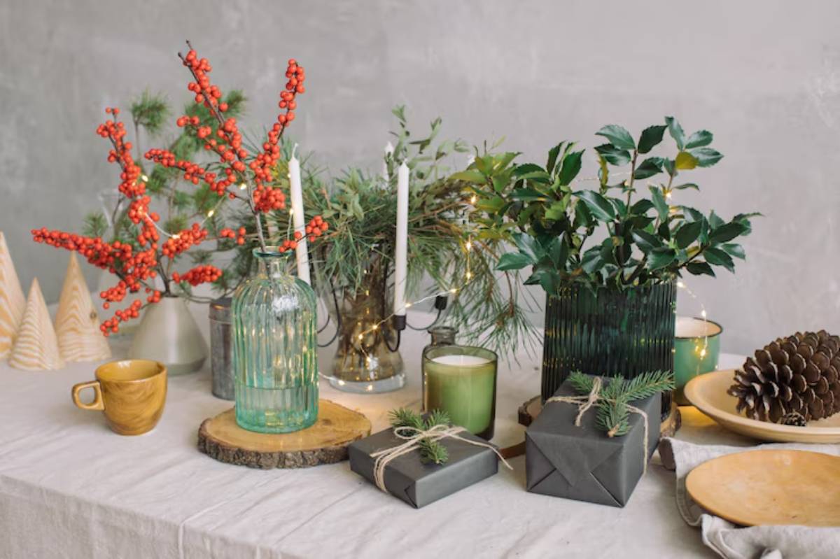 A festive holiday table setup featuring candles, greenery, berries, wrapped gifts, and wooden accents against a textured background.