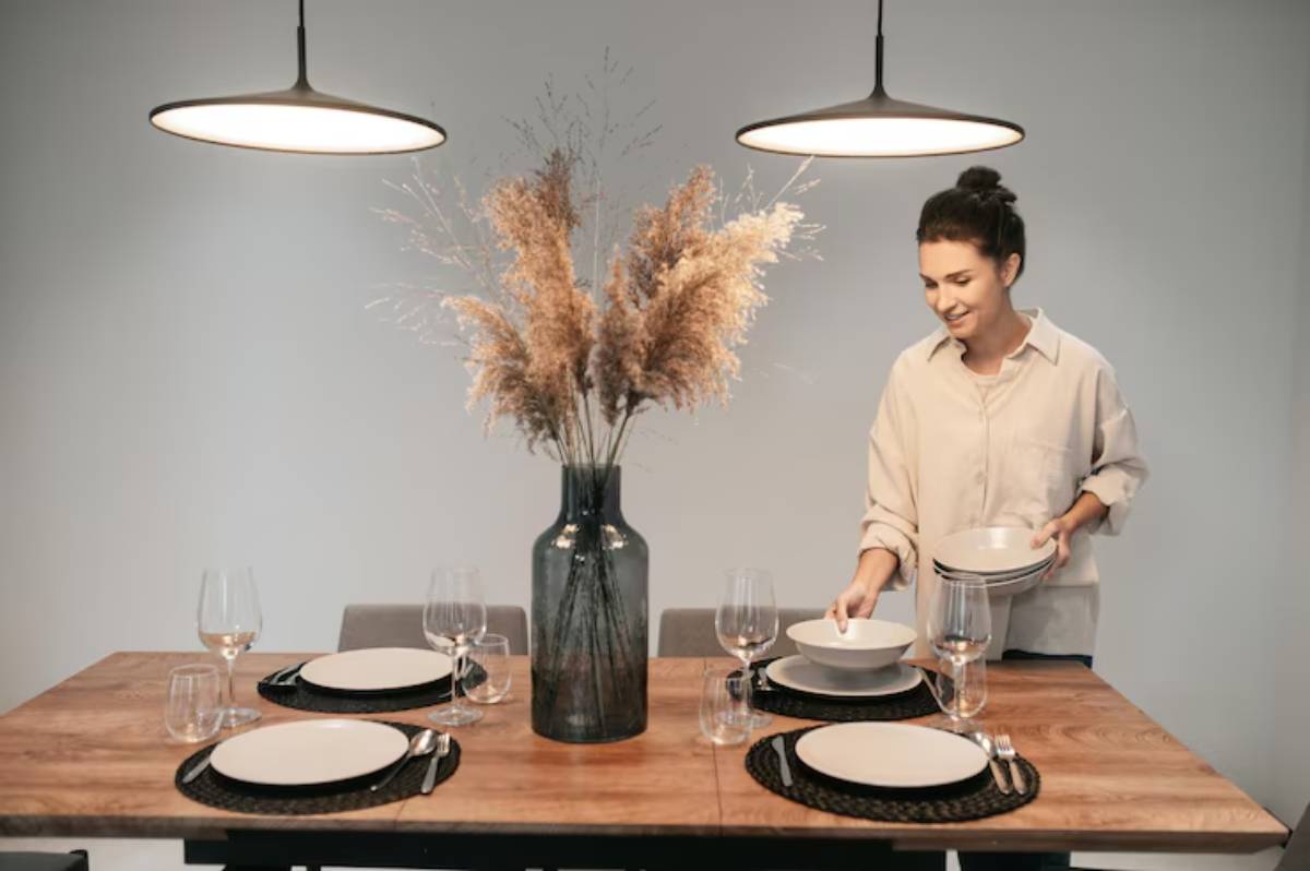 A modern dining table set for four, adorned with elegant glassware, simple white plates, and a vase of dried flowers. Soft lighting overhead.