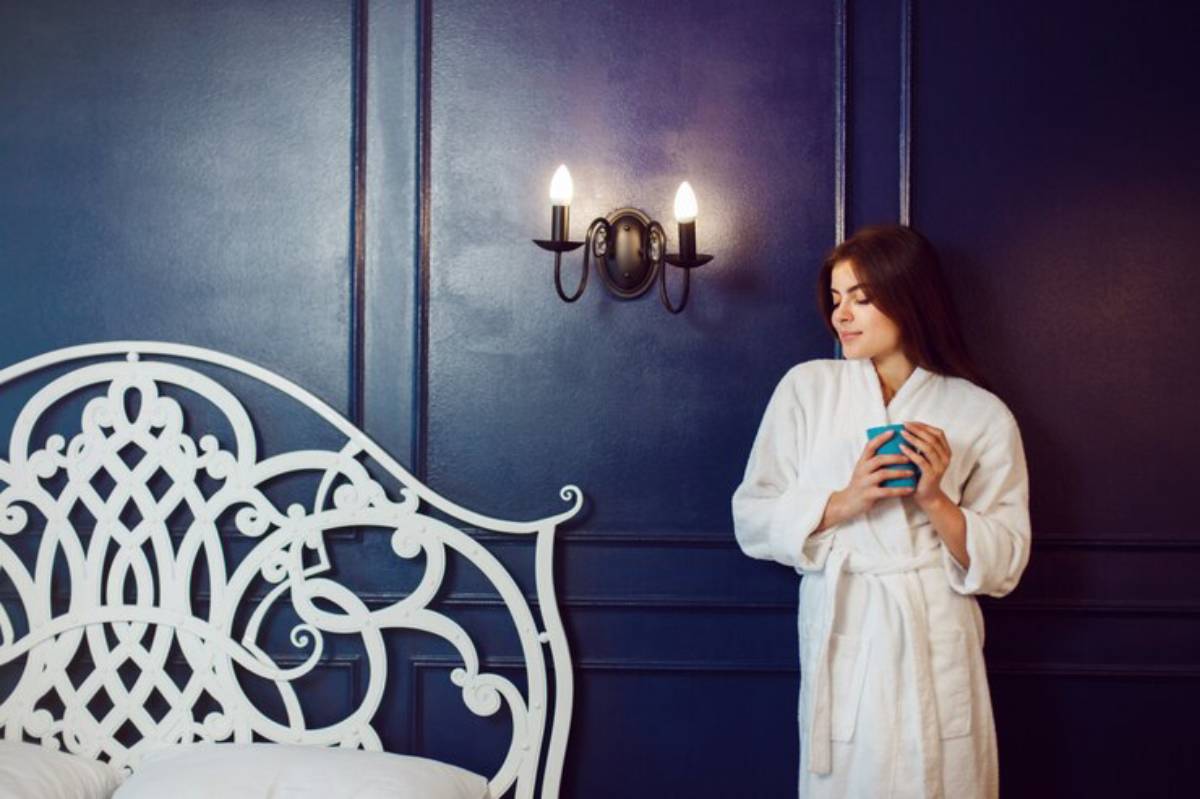 A person in a white bathrobe stands beside a decorative white bed, holding a blue cup against a dark blue wall.