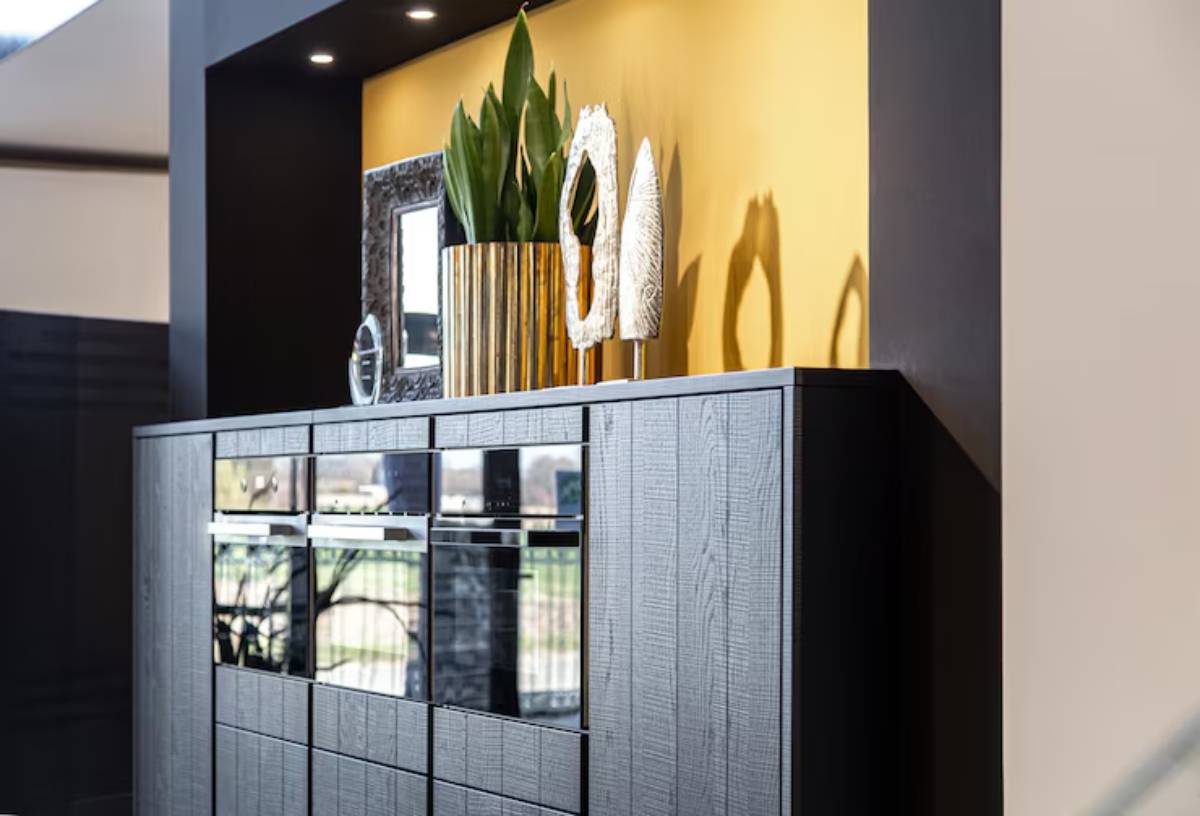 A modern dark wood cabinet with mirrored drawers, adorned with a tall gold vase, decorative sculptures, and a framed mirror against a yellow wall.