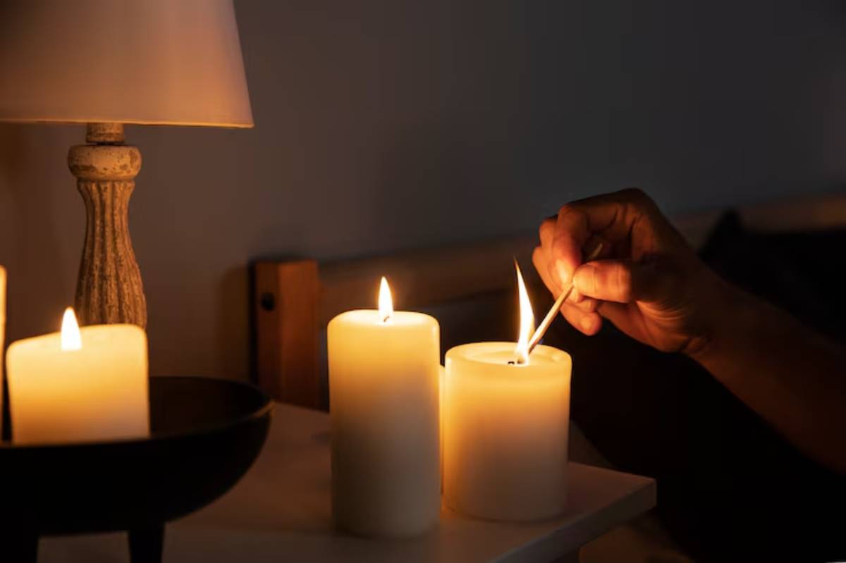 A hand lighting a candle with a matchstick beside two glowing candles on a nightstand, illuminated by soft lamp light.