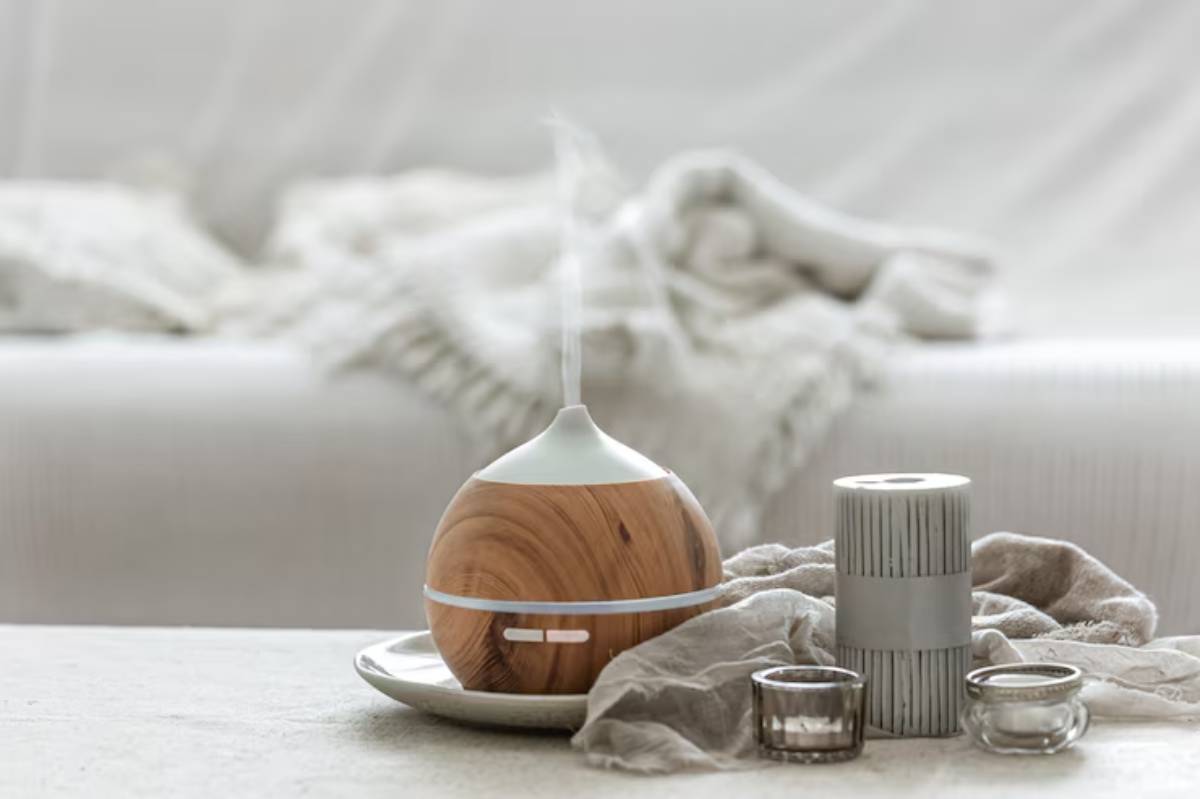 A wooden essential oil diffuser emits steam, accompanied by candles and a gray decorative object on a soft tablecloth in a cozy setting.