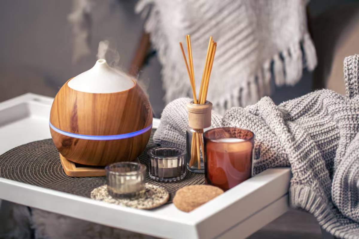 A wooden essential oil diffuser emits steam on a cozy tray with candles, stones, and a soft knit blanket, creating a calming ambiance.