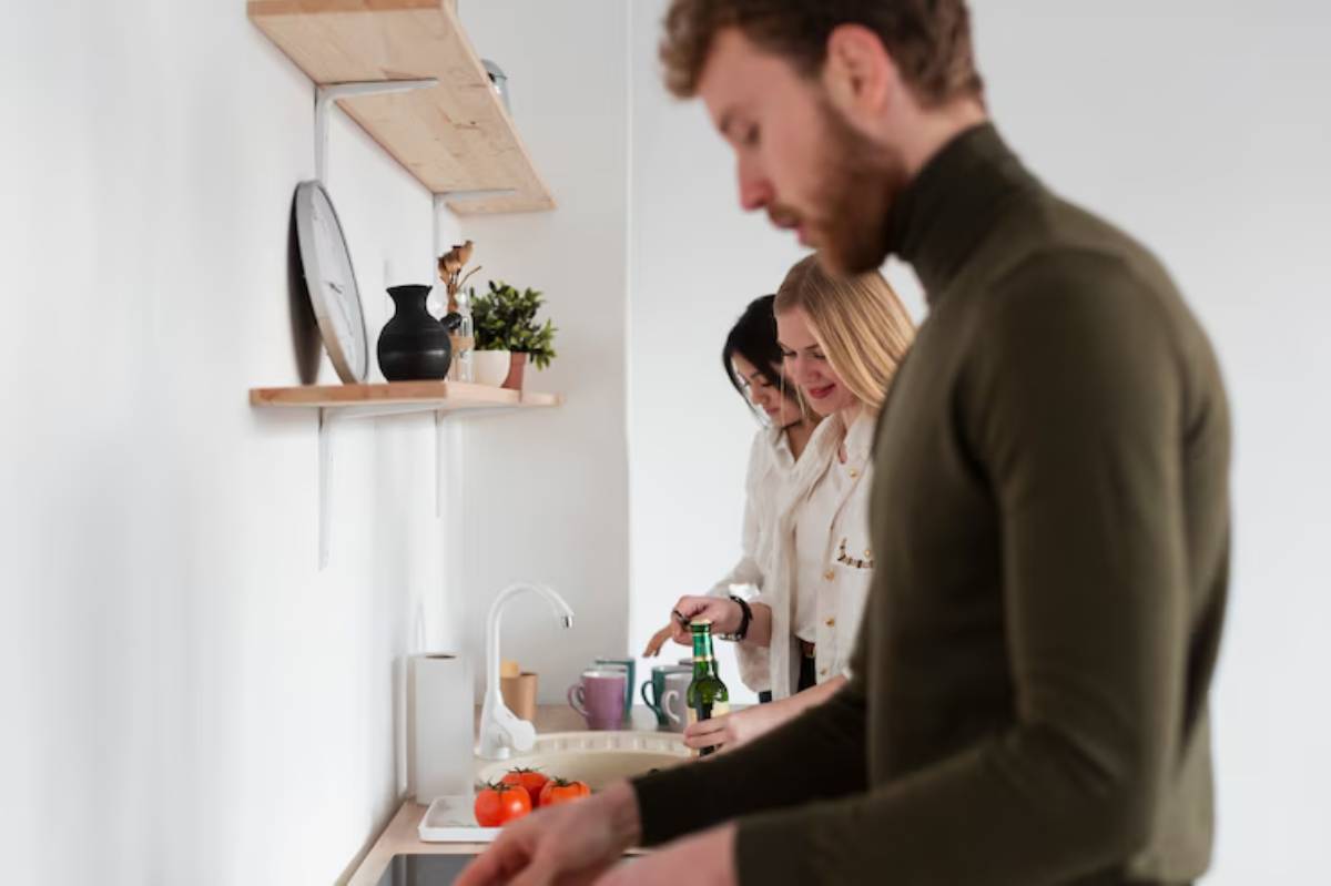 Three individuals are engaged in kitchen activities, preparing food beside a counter with fresh tomatoes and stylish decorations on shelves.