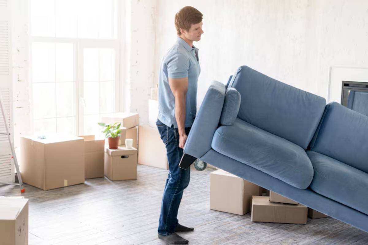 A person lifts a blue couch in a bright, spacious room surrounded by cardboard boxes and a potted plant.