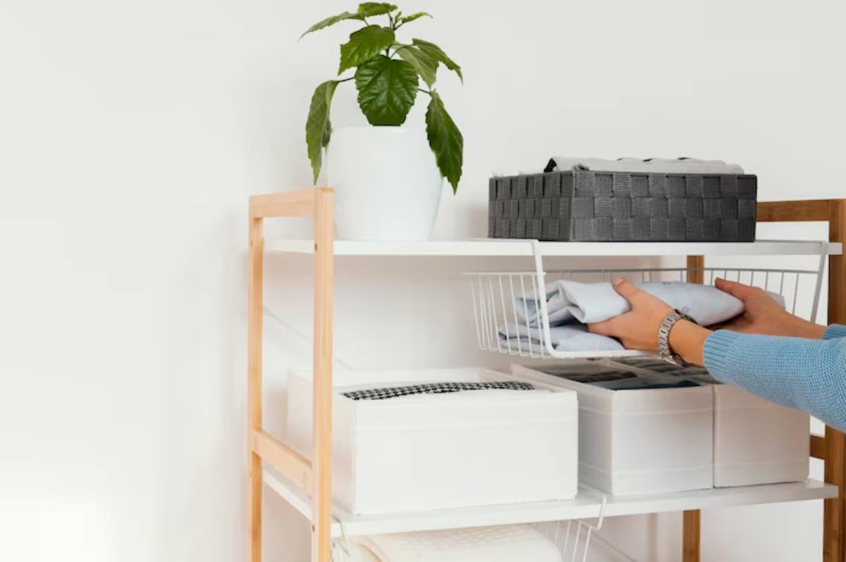 A person in a blue sweater organizes folded clothes in a white wire basket on a wooden shelf, with plant and storage boxes nearby.