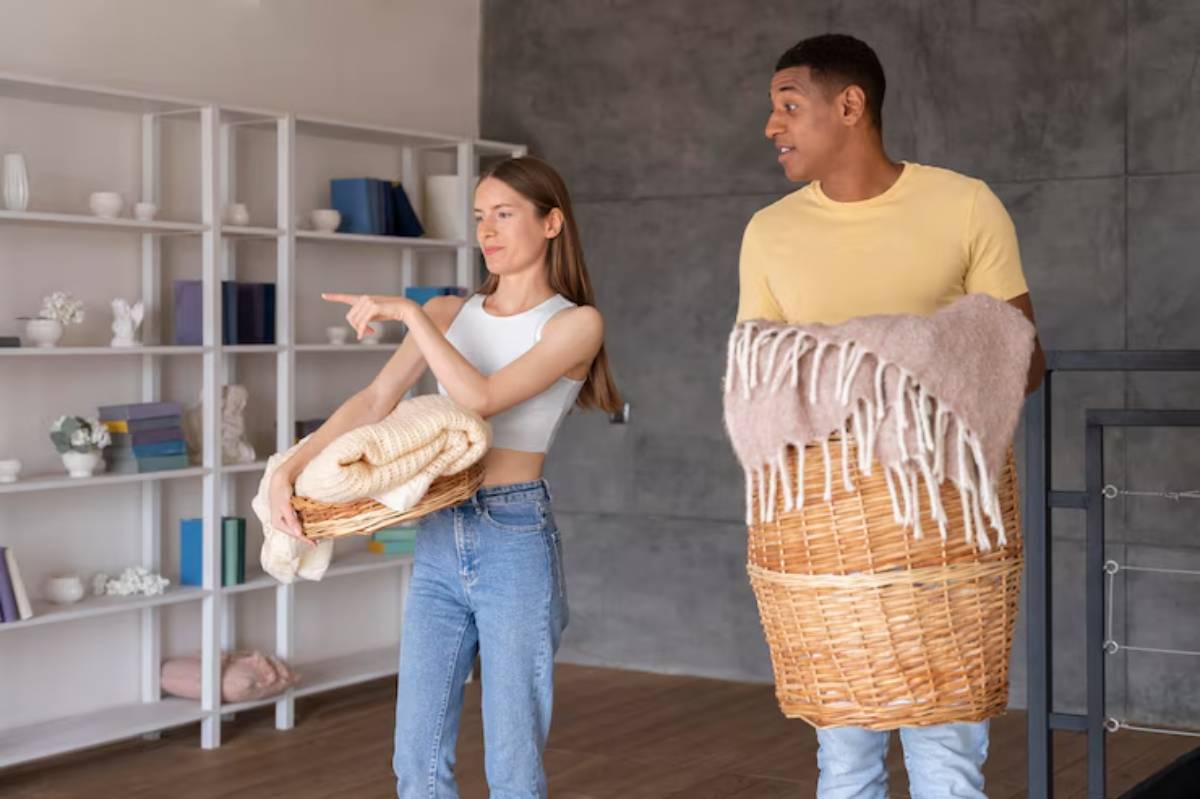 A young woman in a white crop top holds a basket of cream blankets while gesturing, alongside a man carrying a basket with a pink throw.