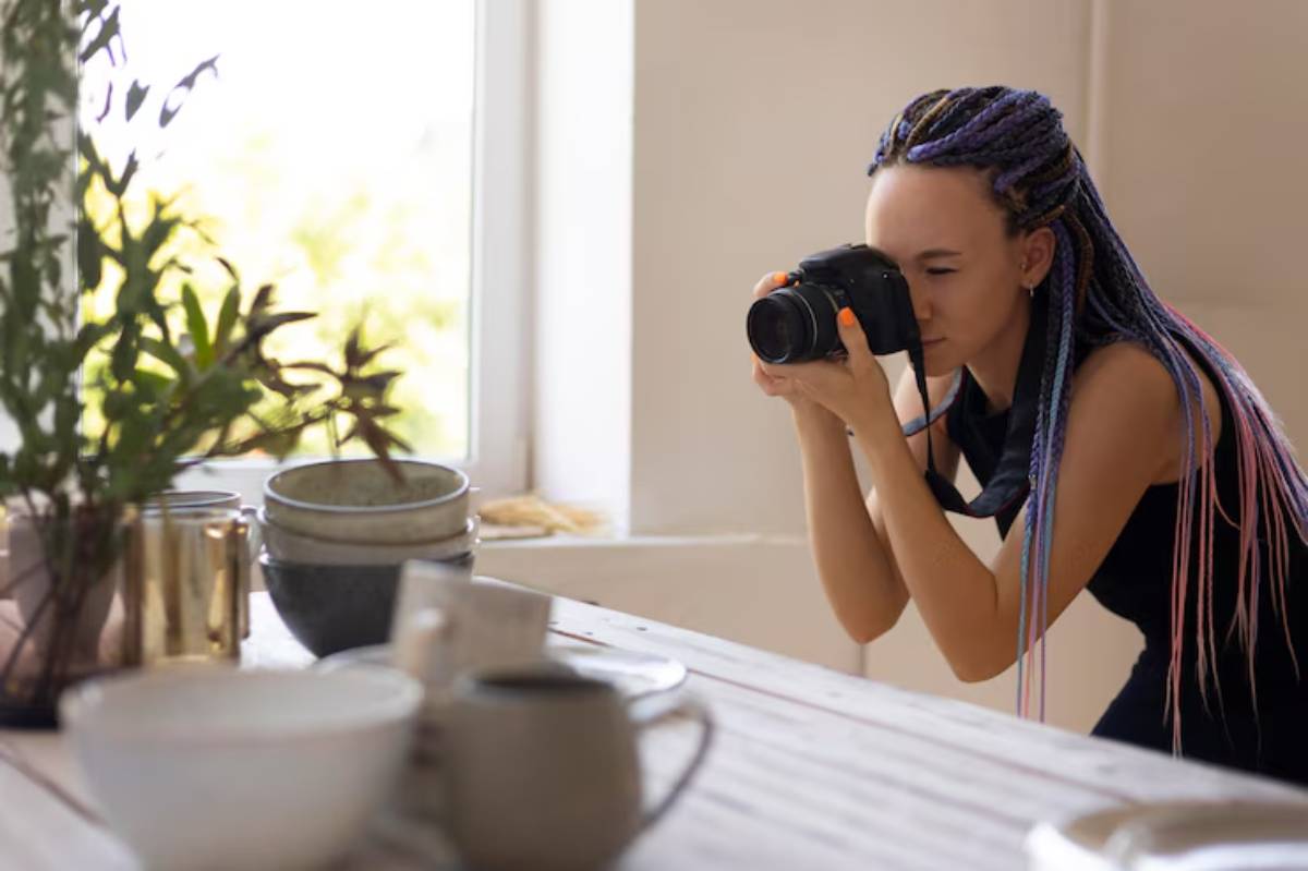 A person with long braided hair is crouched, holding a camera, and focusing on various pottery and plants on a table.