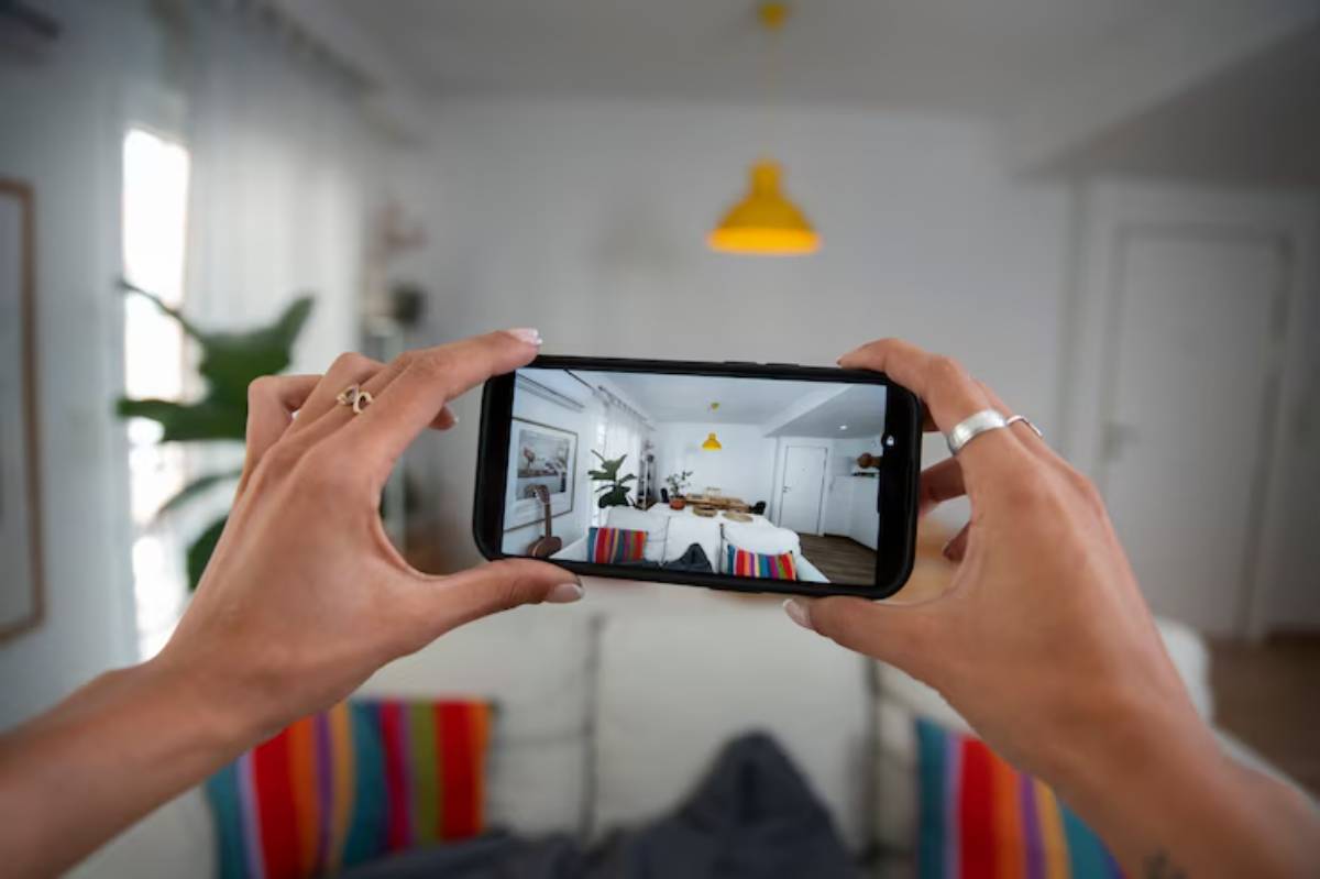 A person holds a smartphone to capture a bright, modern living room featuring colorful cushions and a yellow pendant lamp.