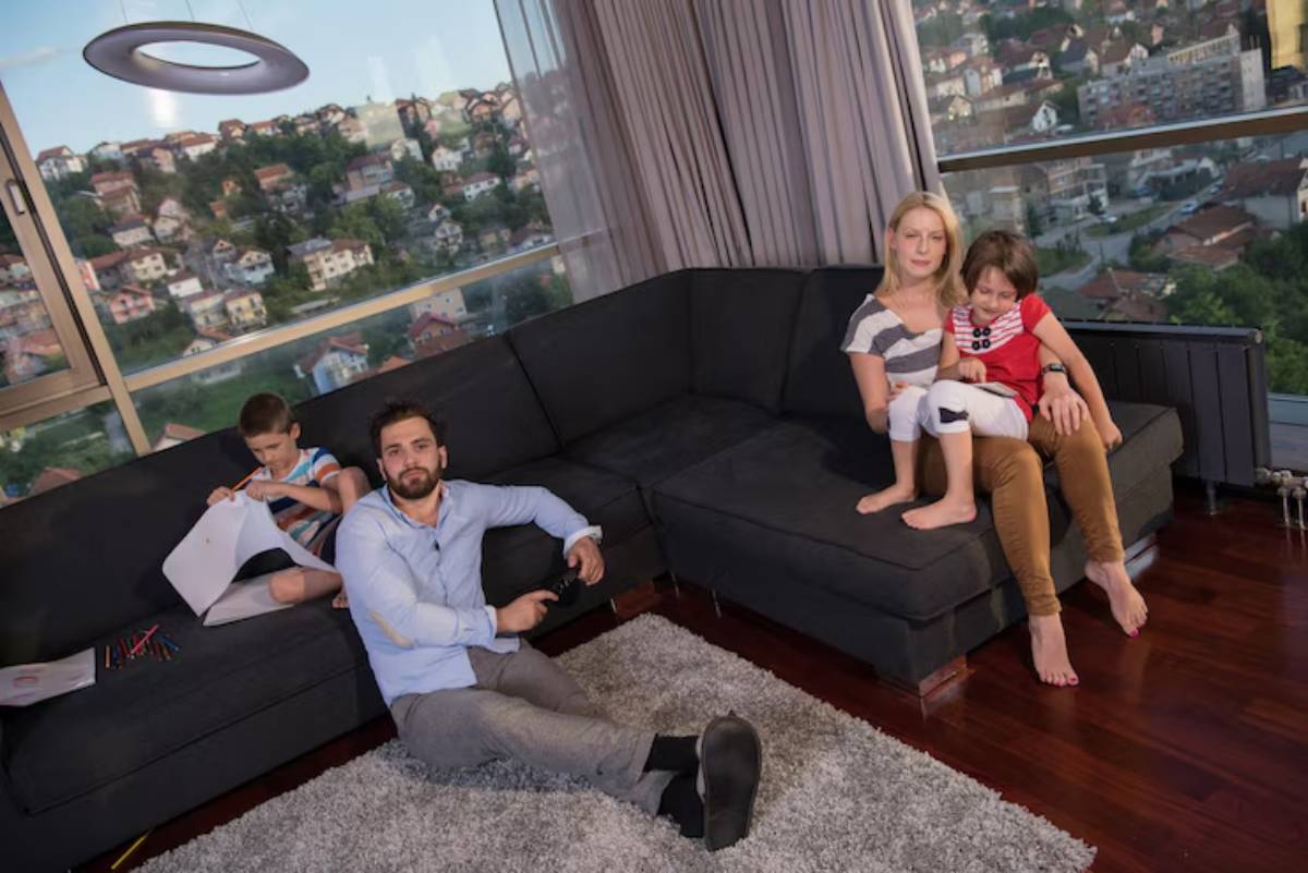A cozy living room scene featuring two children playing, a woman sitting nearby, and a man on the floor, with a city view outside.
