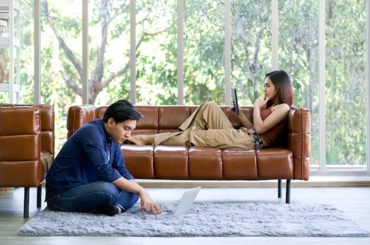 A cozy living room with a brown leather sofa and a person working on a laptop while another relaxes, surrounded by large windows.