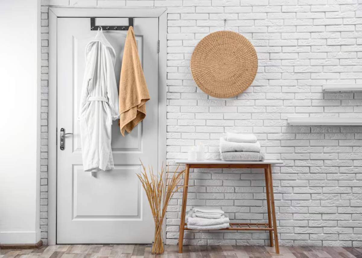 Cozy bathroom corner with a white robe and orange towel hanging on a door, a woven wall decoration, and neatly stacked towels on a table.