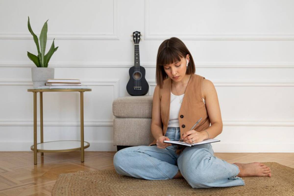 A young woman sits cross-legged on a rug, writing in a notebook, with a potted plant and a ukulele in the background.