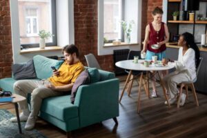 A cozy living room with a teal sofa, a round dining table, and two people preparing drinks, surrounded by warm brick walls and natural light.