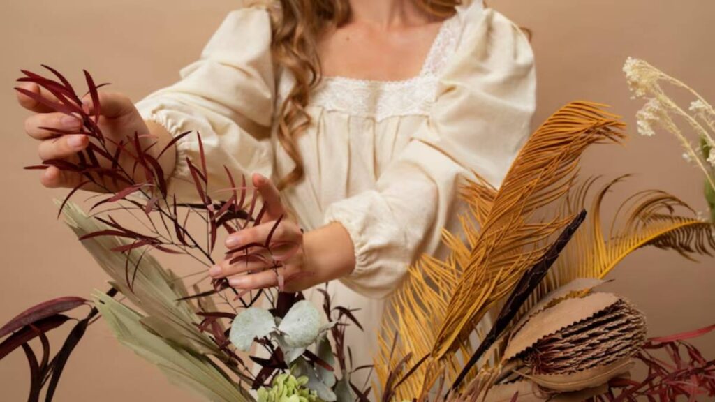A woman in a light cream blouse gently arranges vibrant dried foliage against a soft beige background.