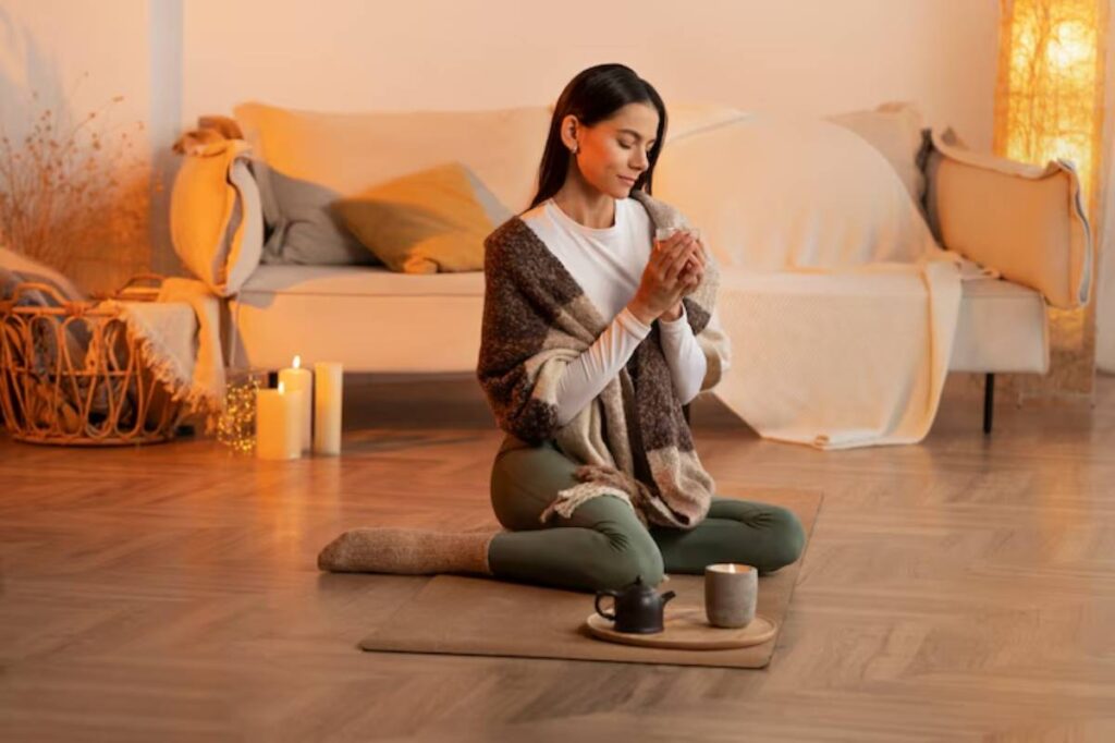 A person sits cross-legged on a mat, holding a cup, surrounded by candles and a teapot, in a cozy, warmly lit living space.