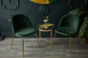 Two elegant green velvet chairs with gold legs flank a glass-topped table, adorned with a decorative flamingo and tropical leaves.