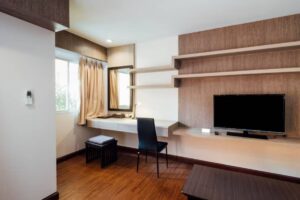 A modern workspace featuring a desk with a chair, a mirror, shelves, and a TV against a wooden accent wall. Natural light filters in.
