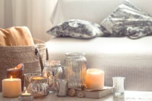 A cozy arrangement of lit candles in various glass holders, on a table next to a soft sofa with patterned pillows.