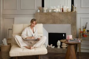 A person in a light robe sits cross-legged on a cream-colored sofa, writing in a notebook beside a stylish fireplace and decorative items.