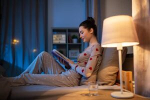A person relaxing in bed, wearing heart-patterned pajamas, reading a book beside a soft glowing lamp and a glass of water.