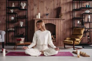 A person in white attire meditates on a purple mat in a cozy, wooden room with candles, a fireplace, and decorative shelves.