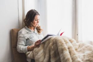 A woman sits comfortably in a chair, reading a book while wrapped in a soft blanket, with sunlight streaming through sheer curtains.