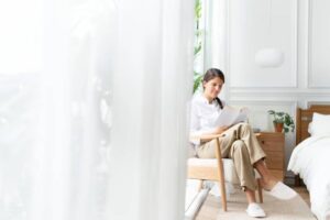 A person sits in a cozy chair reading a book, surrounded by light and greenery in a modern, bright room.