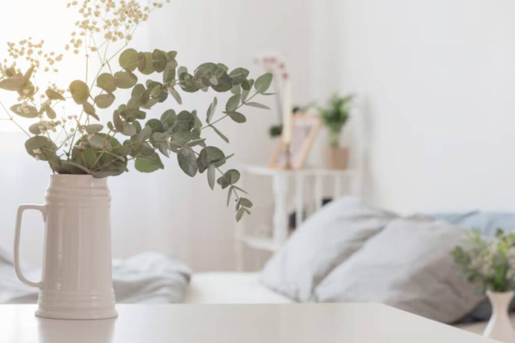 A white vase with greenery sits on a table, with soft pillows and bright natural light in a cozy, minimalistic room.