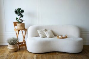 A curved white sofa with two cushions, a wooden folding chair holding a potted plant, and a woven basket with flowers, in a minimalistic room.
