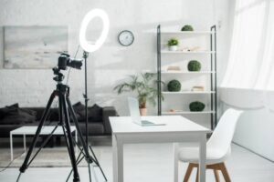 A bright, minimal workspace featuring a camera on a tripod, a ring light, a laptop, and a white chair, with plants in background.