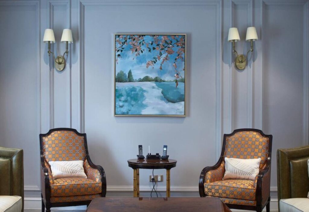 Cozy sitting area featuring two patterned chairs, a wooden table with phones, and a vibrant landscape painting on a soft blue wall.