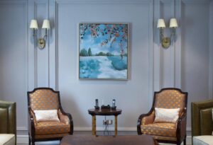 Cozy sitting area featuring two patterned chairs, a wooden table with phones, and a vibrant landscape painting on a soft blue wall.